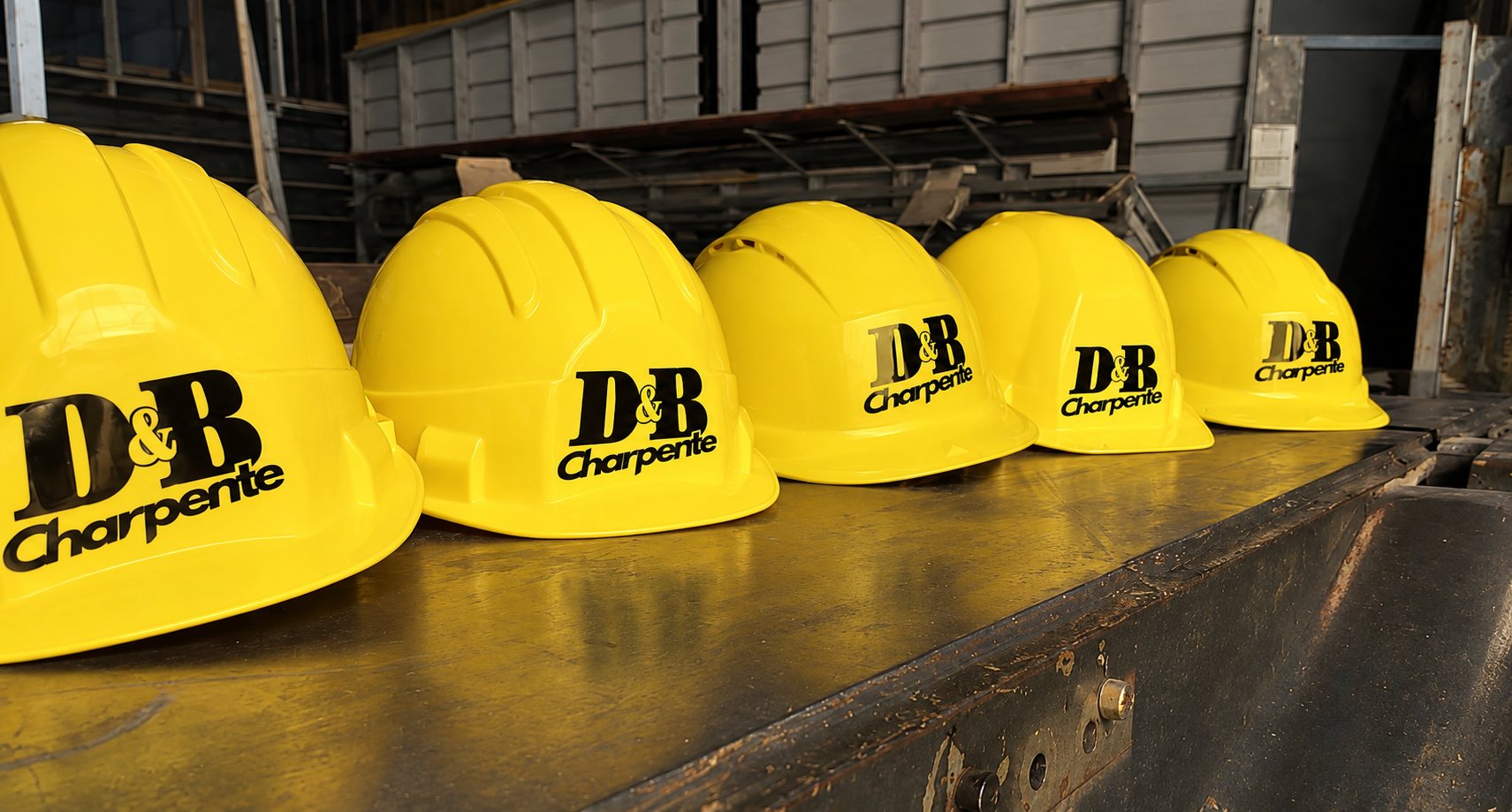 Cinq casques de chantier jaunes portant le logo D&B Charpente sont alignés sur un établi en métal dans un atelier.