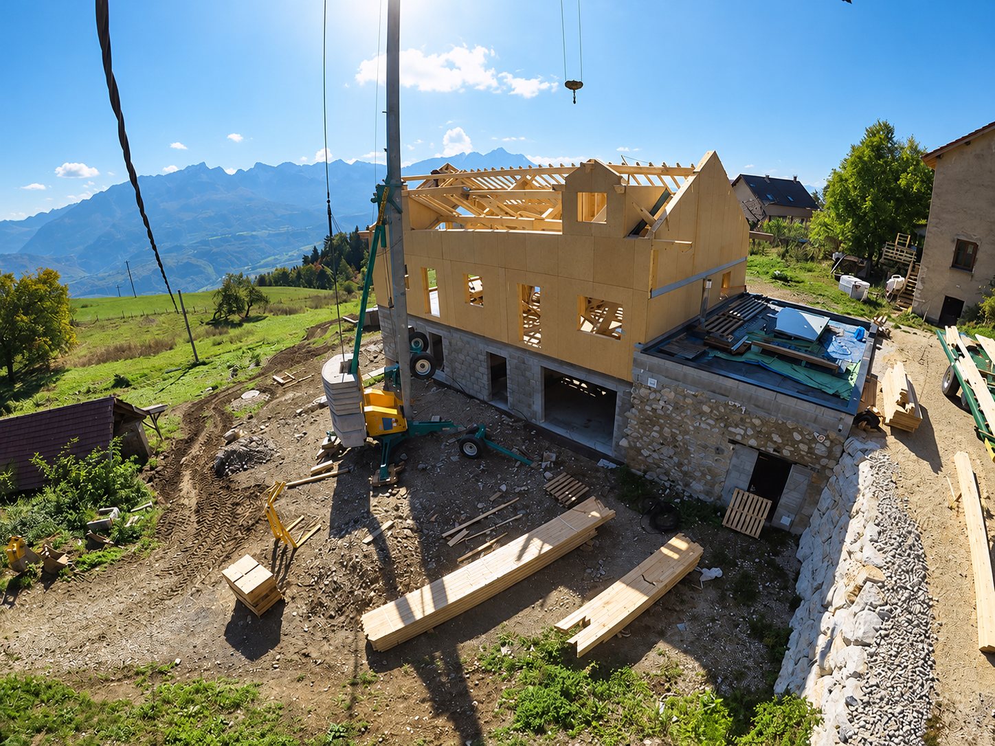 Vue aérienne d'un chantier de maison en panneaux de bois avec charpente en cours de pose, sur un soubassement en pierre, avec une grue et des montagnes en arrière-plan.