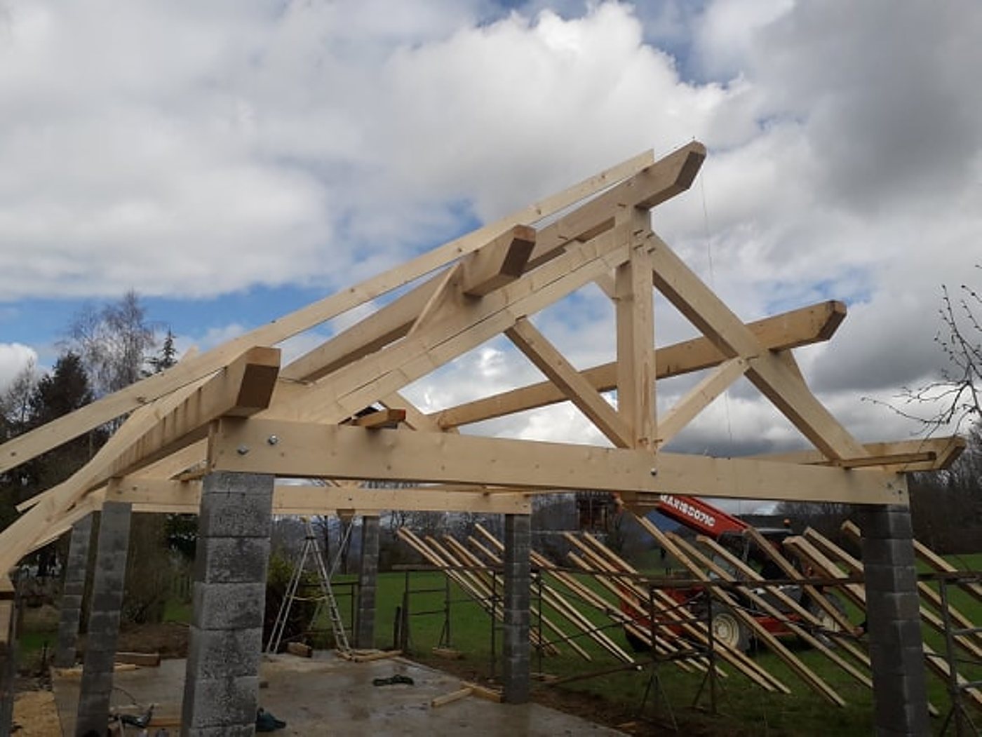 Une charpente traditionnelle en bois en cours de montage sur des piliers en parpaings, avec une grue rouge et des chevrons empilés en arrière-plan sous un ciel nuageux.