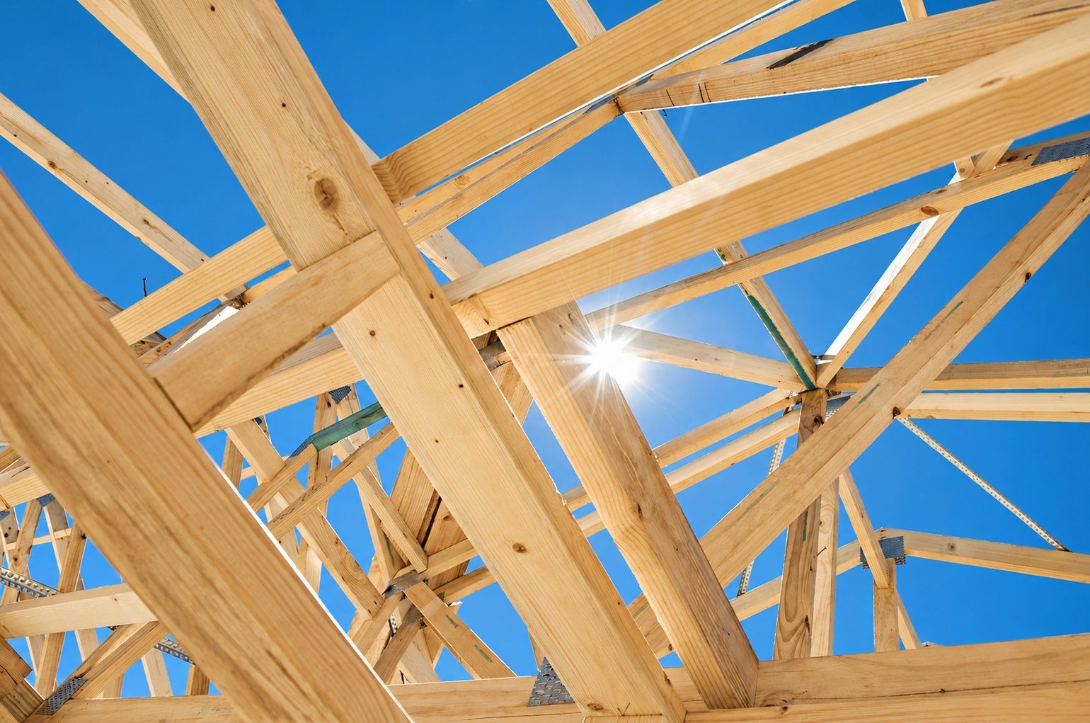 Vue en contre-plongée d'une charpente en bois clair en cours de construction, avec le soleil qui brille à travers les fermes sur un ciel bleu.