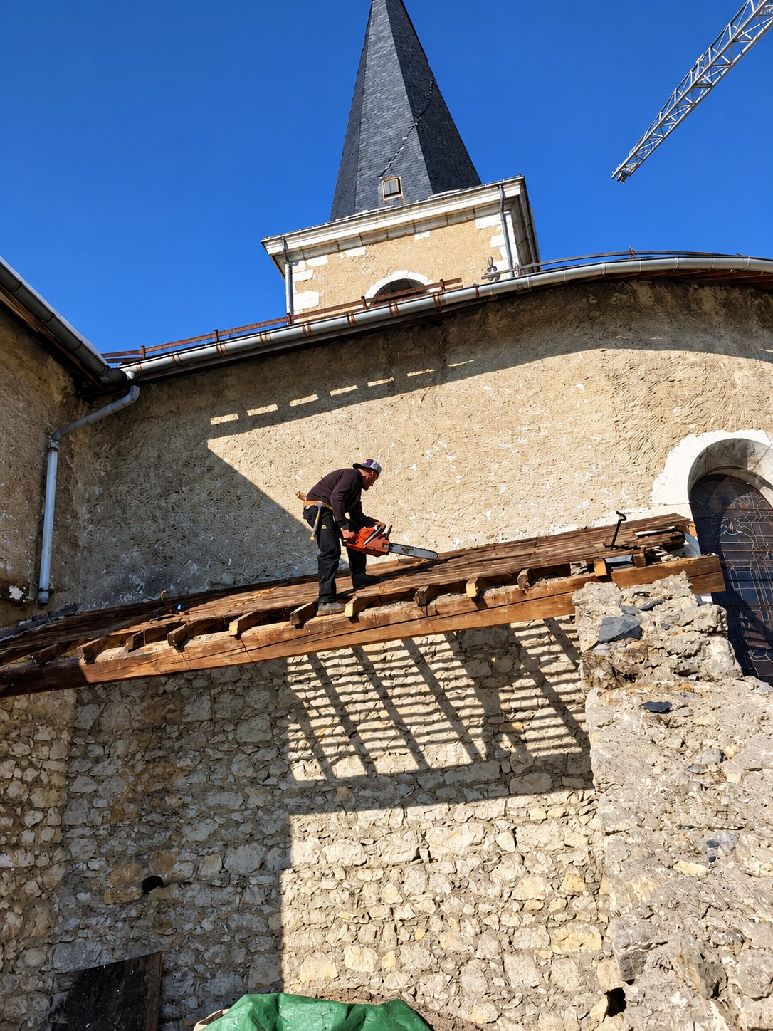 Un ouvrier muni d'une tronçonneuse découpe la charpente en bois d'un toit d'église en travaux, sous un ciel bleu, avec le clocher à ardoises et une grue en arrière-plan.