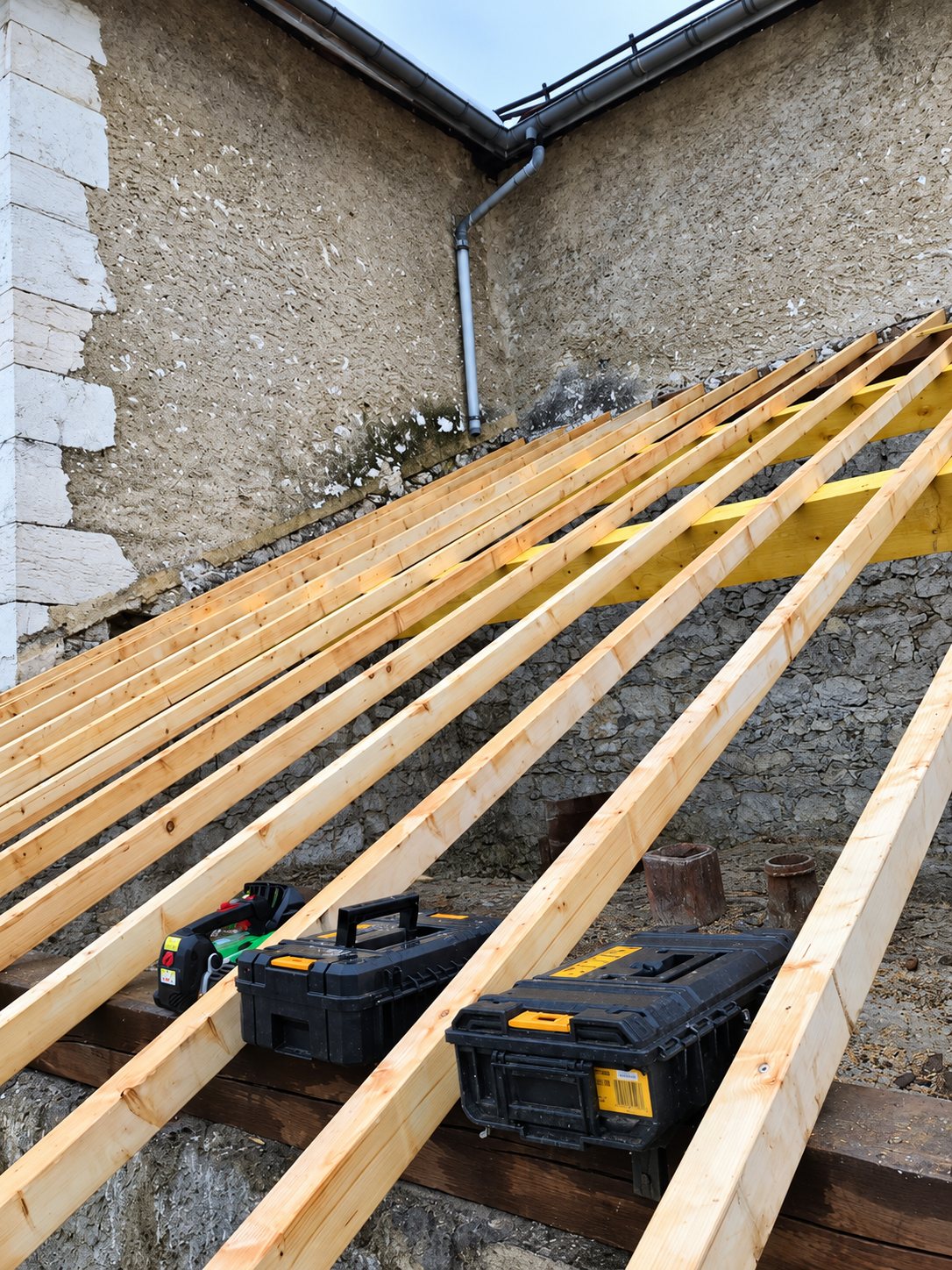 Des chevrons en bois neufs sont posés sur la pente d'un toit contre un mur en pierre, avec deux caisses à outils noires et jaunes posées dessus.