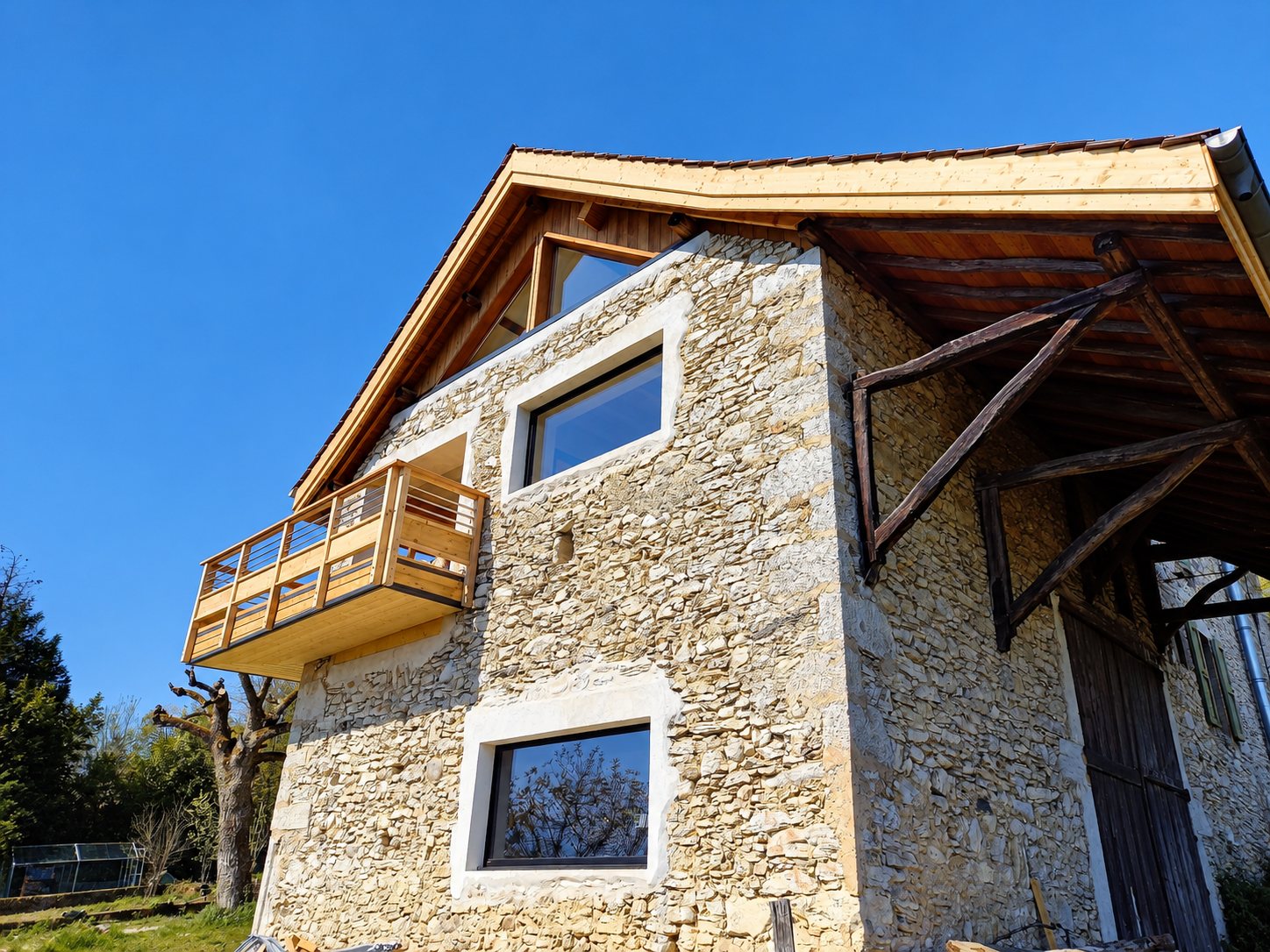 Façade d'une ancienne grange en pierres apparentes rénovée, avec grandes fenêtres, balcon en bois clair et charpente apparente sous un ciel bleu.