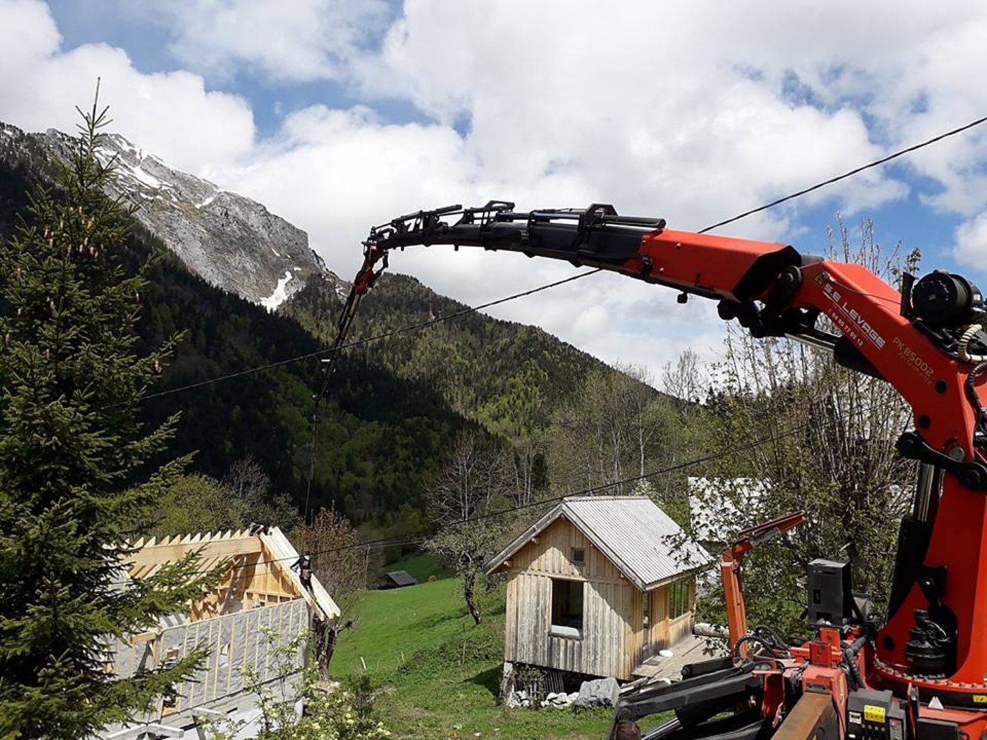 Un bras de grue orange s'étend au-dessus d'une charpente en cours de montage, avec une cabane en bois et des montagnes en arrière-plan.
