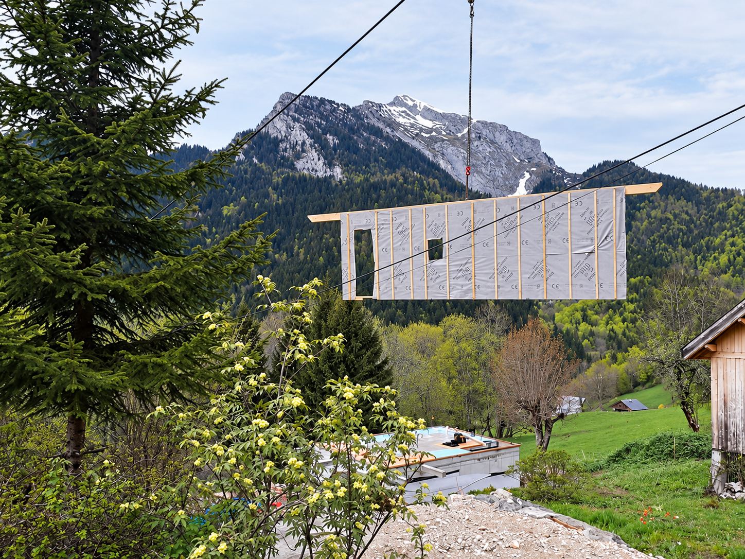 Un panneau de mur à ossature bois recouvert de membrane est suspendu à une grue devant une montagne boisée, au-dessus d'un chantier de construction.