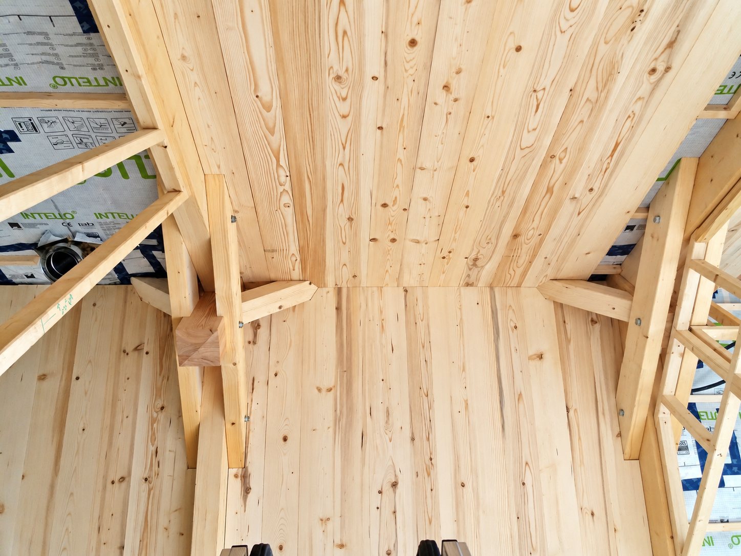 Vue vers le haut d'un plafond en lambris de bois clair posé entre des poutres de charpente, avec une membrane Intello visible sur les côtés.