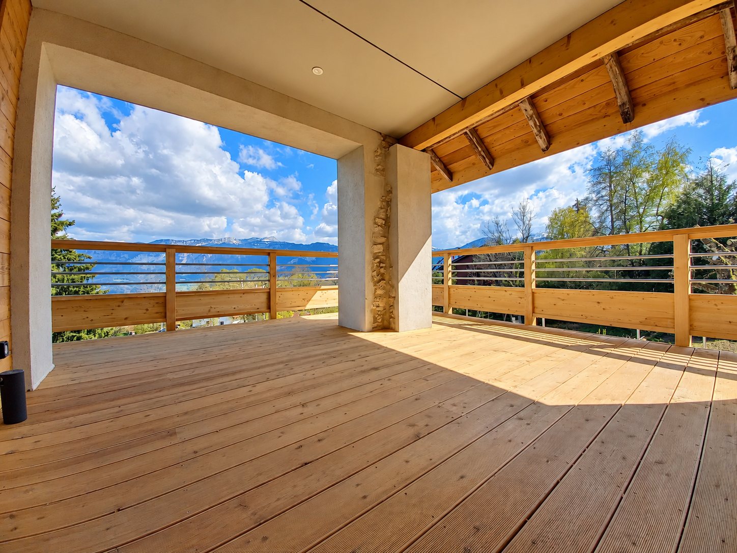 Terrasse en bois neuve et couverte, avec garde-corps à lisses horizontales, ouverte sur un paysage de montagnes et de ciel nuageux.