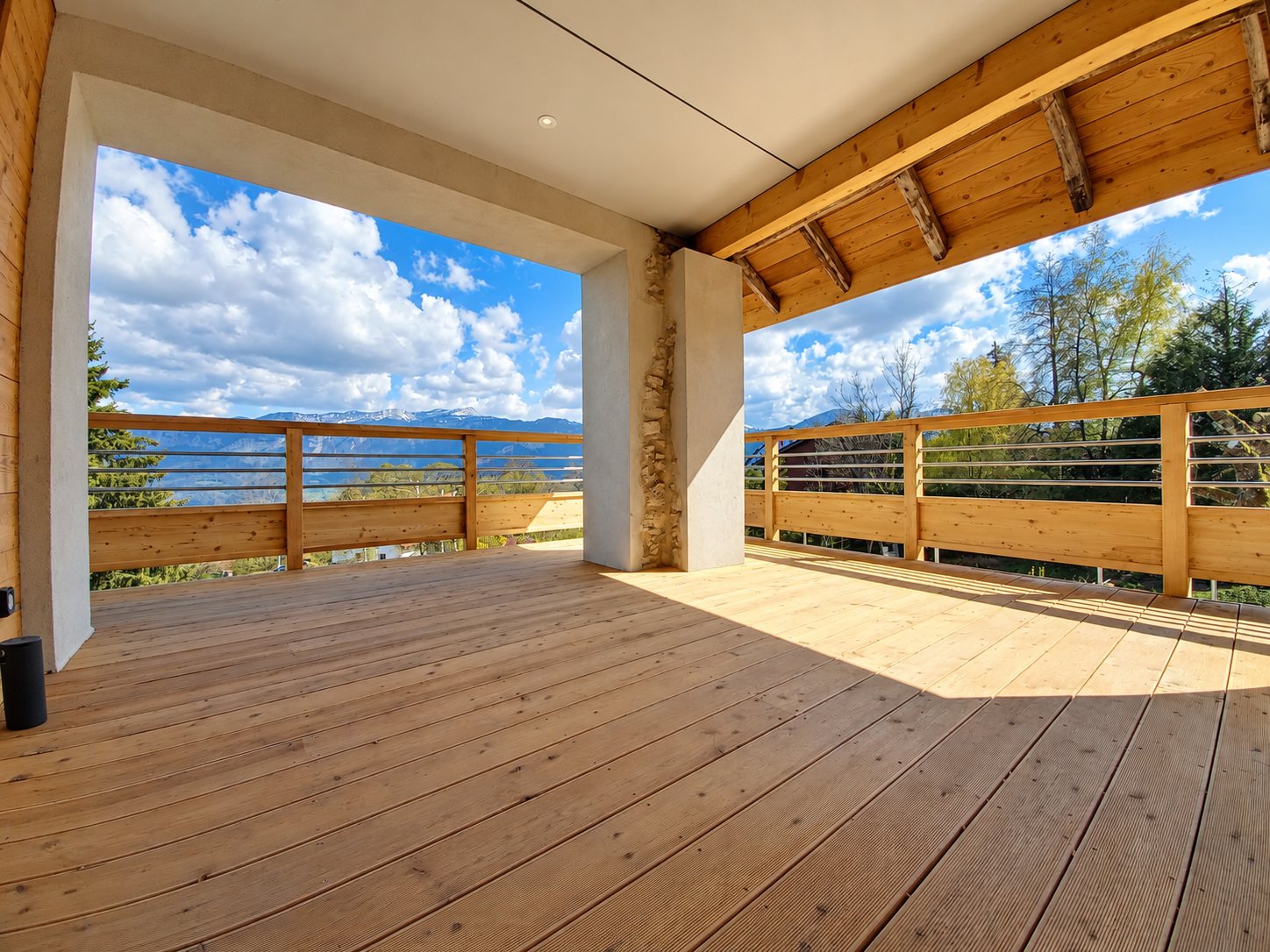 Grande terrasse en bois avec garde-corps à lames horizontales, sous une charpente apparente, offrant une vue dégagée sur les montagnes et le ciel nuageux.