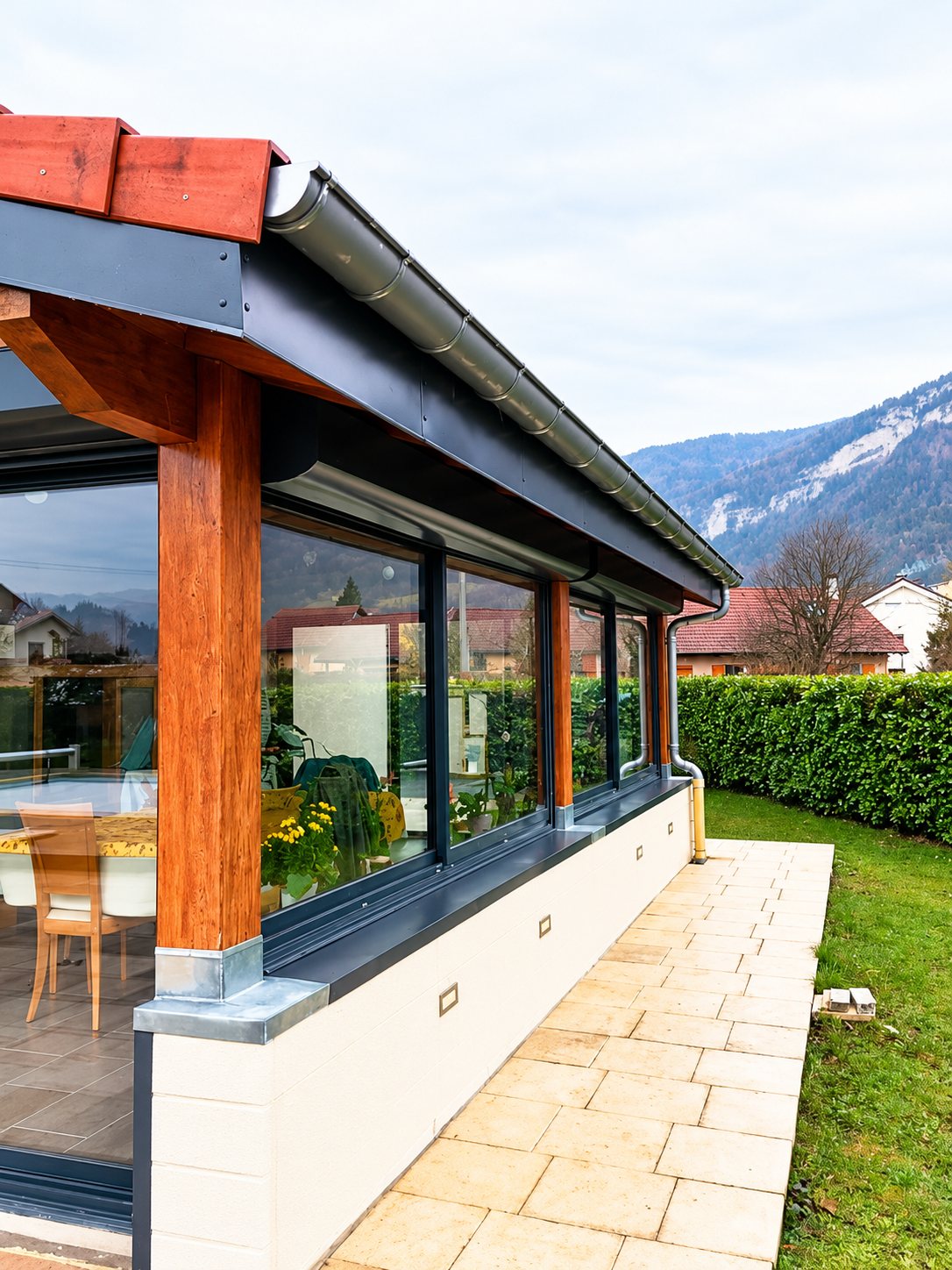Une véranda aux grandes baies vitrées et aux poteaux en bois, bordée d'une terrasse dallée et d'une haie, avec des montagnes en arrière-plan.